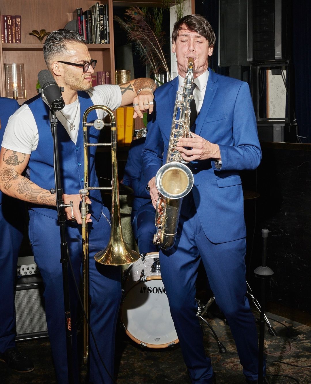 A man in a blue suit plays the saxophone while a trombone player watches on